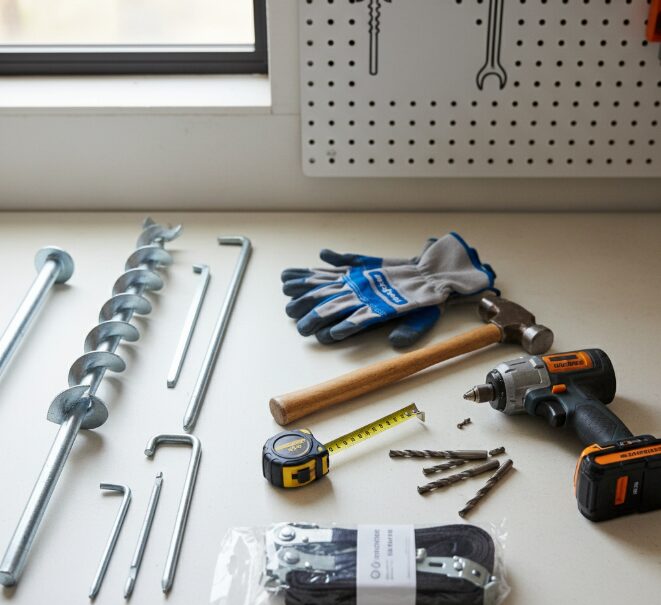 Various tools and materials are neatly arranged: measuring tape, gloves, hammer, drill bits, different types of ground anchors (spiral anchors, J-anchors), and tying strap kits.