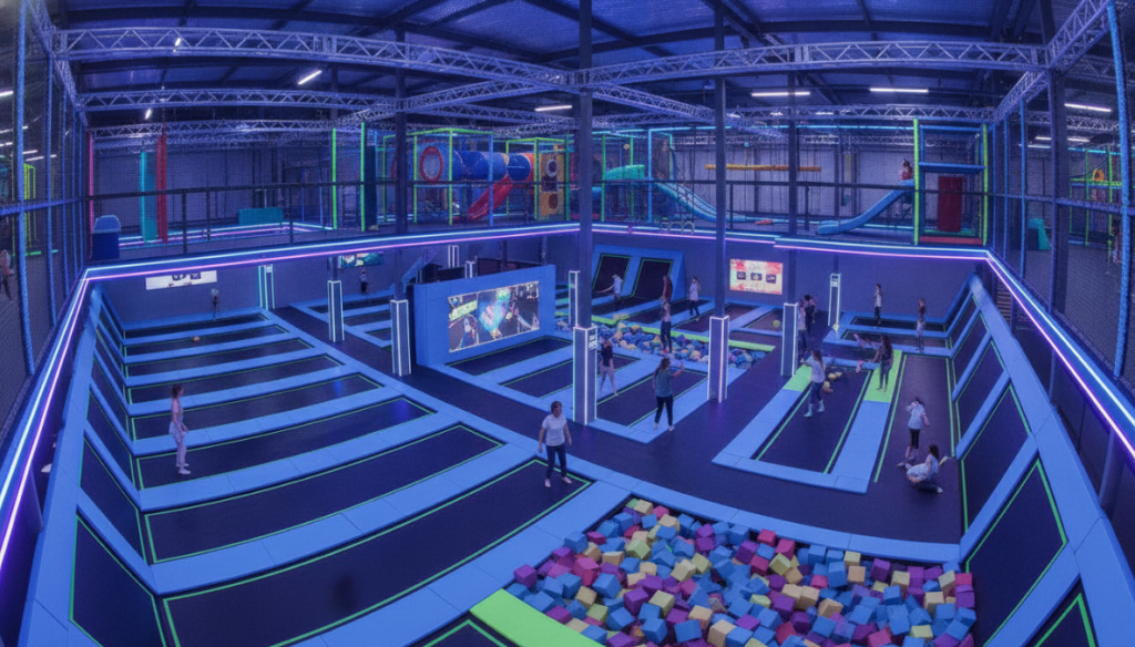 Wide-angle view of a custom multi-level indoor trampoline park featuring neon lighting, interconnected jump zones, a colorful foam pit, and an elevated soft play structure with clear crowd flow pathways